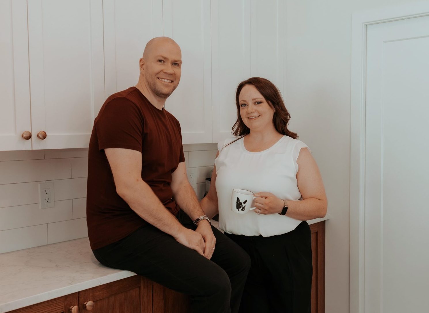 Two realtors posing in a modern kitchen. The man, Shawn, casually sitting on a wooden countertop with white upper cabinets behind him, is wearing a maroon shirt and black pants. The woman, Sarah, standing next to him holding a mug with a Boston Terrier dog design, is dressed in a white blouse and black pants. Both are smiling, exuding warmth and approachability in this contemporary, well-lit space.