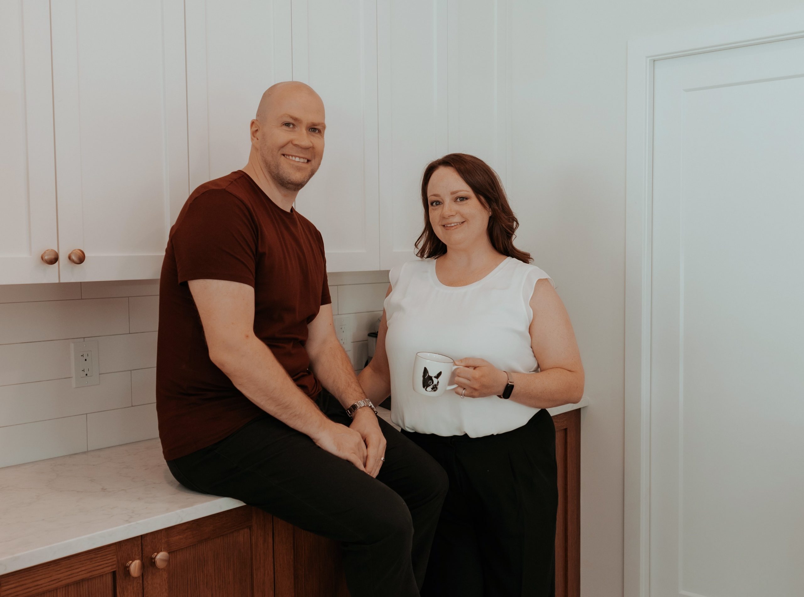 Two realtors posing in a modern kitchen. The man, Shawn, casually sitting on a wooden countertop with white upper cabinets behind him, is wearing a maroon shirt and black pants. The woman, Sarah, standing next to him holding a mug with a Boston Terrier dog design, is dressed in a white blouse and black pants. Both are smiling, exuding warmth and approachability in this contemporary, well-lit space.