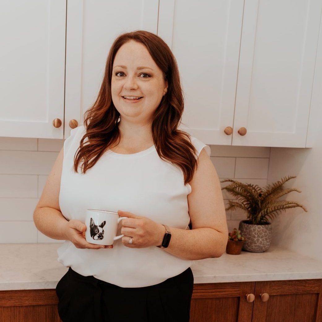 A female realtor, Sarah, standing in a modern kitchen, holding a mug with a Boston Terrier dog design. She is wearing a white blouse and black pants, smiling warmly at the camera. The kitchen features white upper cabinets and wooden lower cabinets with brass knobs, alongside a small decorative plant, creating a clean and inviting space.