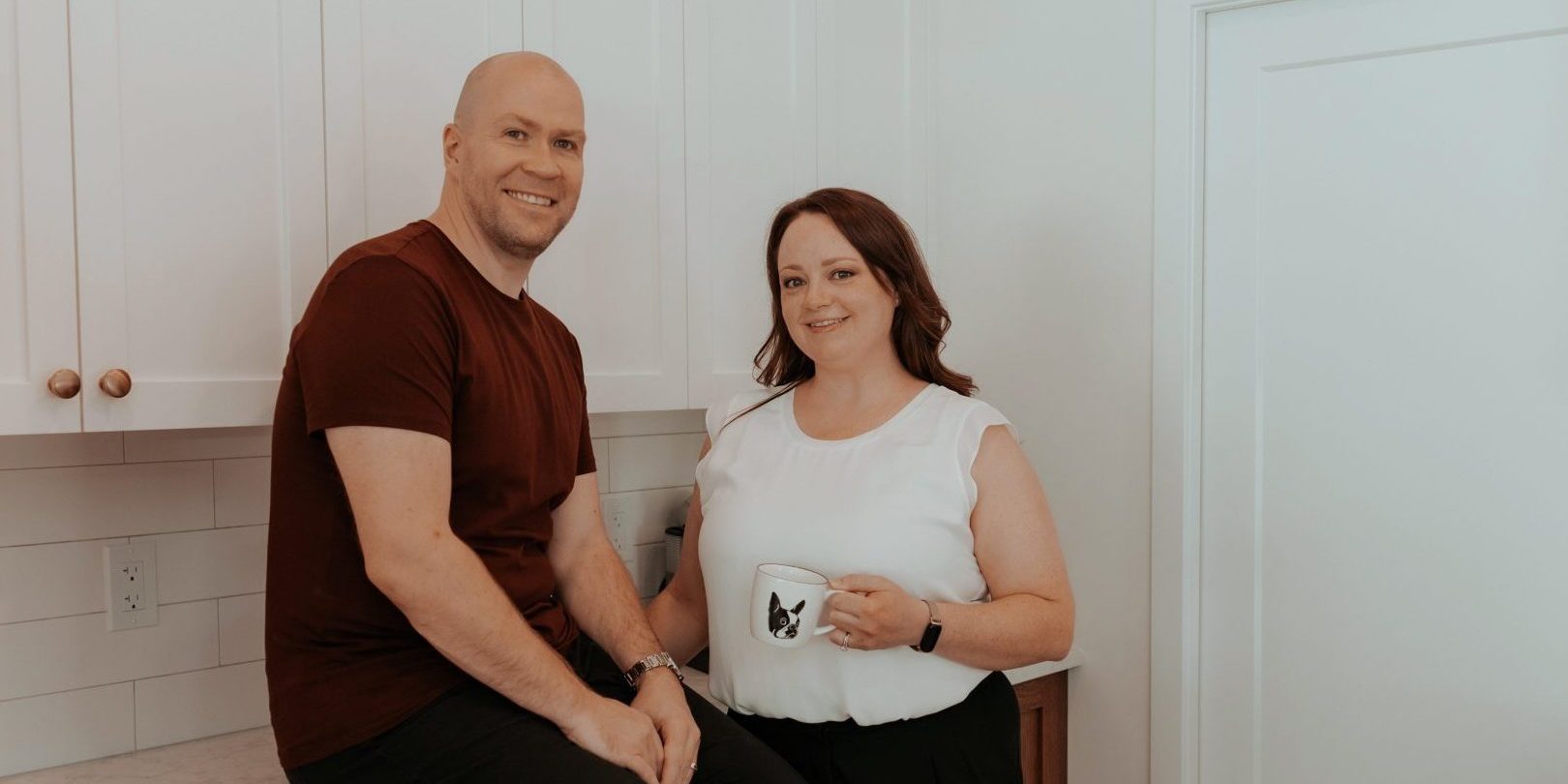 Two realtors posing in a modern kitchen. The man, Shawn, casually sitting on a wooden countertop with white upper cabinets behind him, is wearing a maroon shirt and black pants. The woman, Sarah, standing next to him holding a mug with a Boston Terrier dog design, is dressed in a white blouse and black pants. Both are smiling, exuding warmth and approachability in this contemporary, well-lit space.